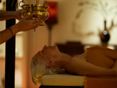 Woman lies on a massage bed in an orange towel as a therapist pours oil over her forehead from a gold bowl