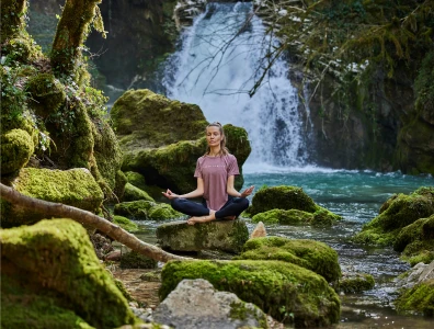 Woman sitting in lotus position on rocks meditating by a waterfall 