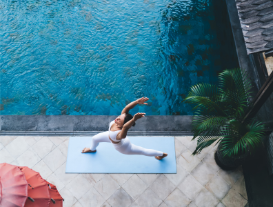 Woman in white activewear practices yoga on a blue mat next to a swimming pool
