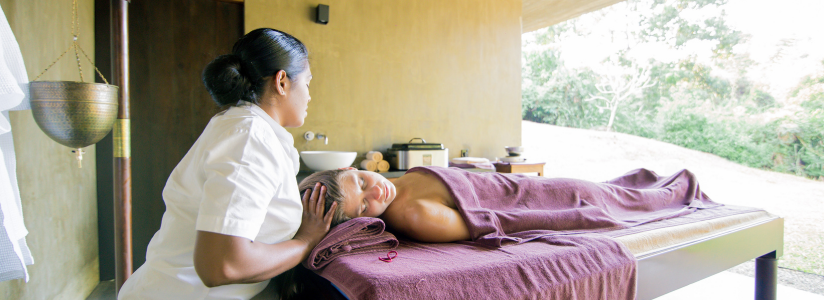 Ayurveda healing Woman lies on a massage bed in a purple towel as an Ayurveda therapist lays hands on her head