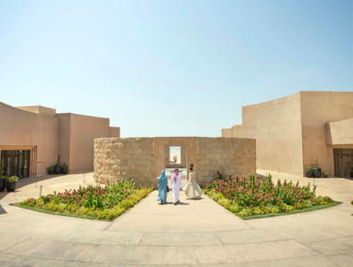 Low-rise sand-coloured resort with circular-like buildings, with gentle flowers and three women walking towards an open door