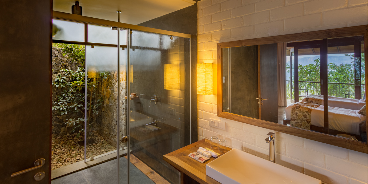 Glass-walled shower showing gardens outside, a vanity unit with a large white sink and a mirror on the wall