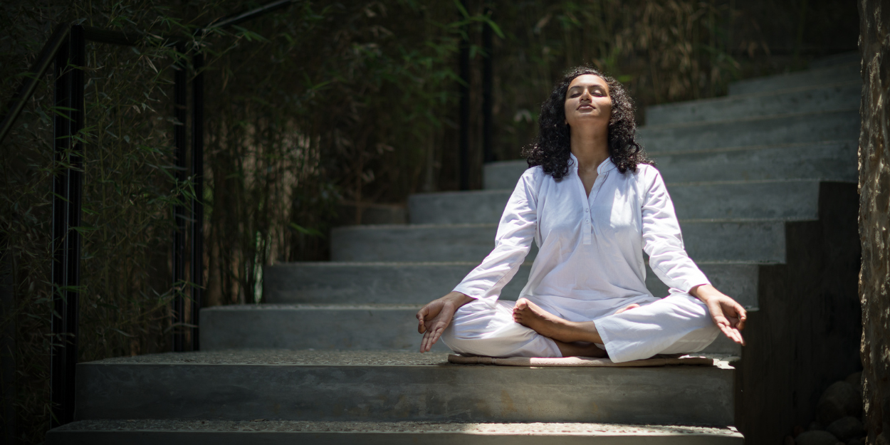 Woman in white sits cross legged with her eyes closed and palms up on a stone staircase