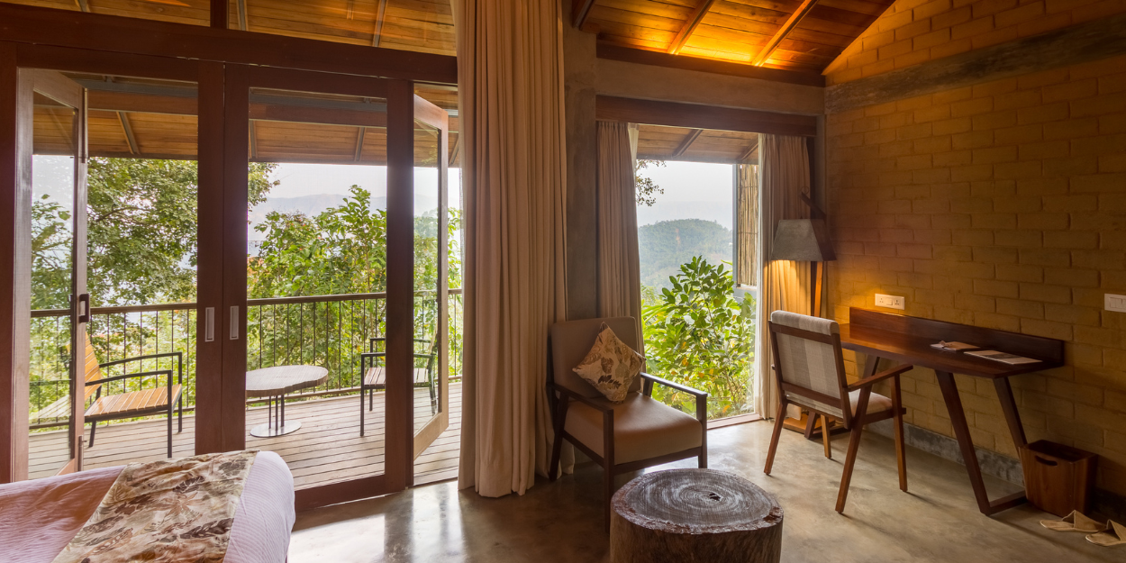 Hotel room with neutral colouring, desk space, a chair and coffee table and floor-to-ceiling patio doors opening onto a furnished terrace