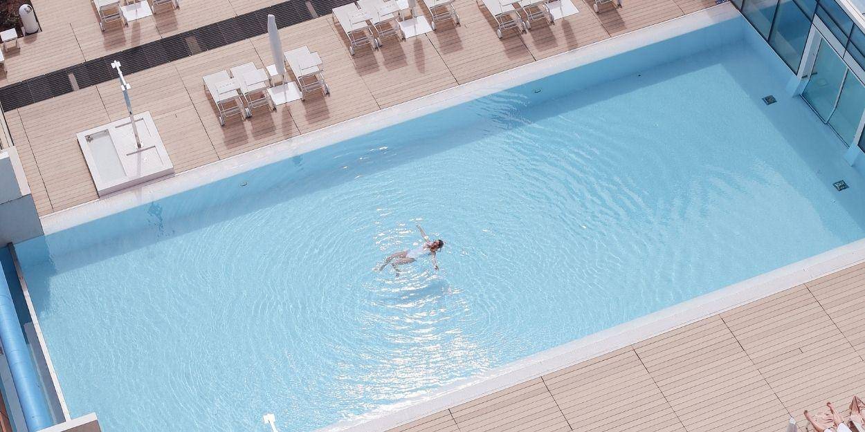 Woman in a white swimsuit floats on her back in a rectangular pool surrounded by wooden decking and loungers