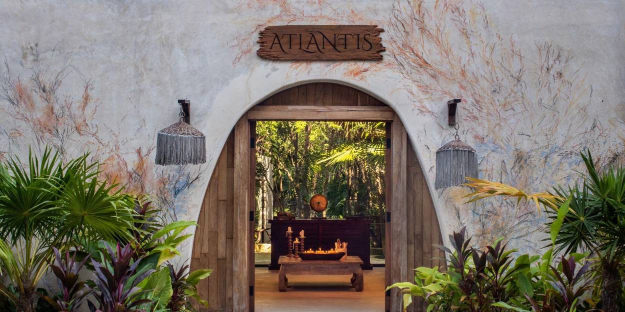 Spa at Palmaïa – The House of AïA Stone-walled entrance to the Atlantis Spa, with an arched entrance fringed by greenery peering through to a wooden table covered in candles