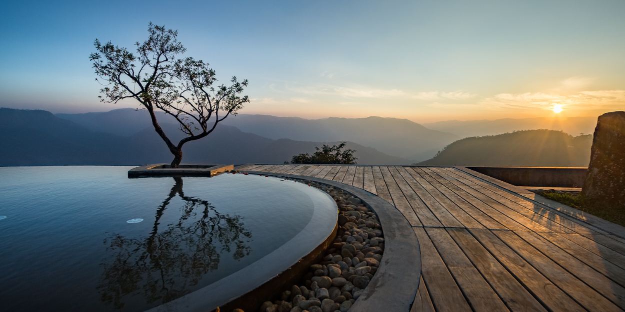 Curved edge of an infinity pool set in wooden decking with a tree next to it and views of misty mountains at sunset