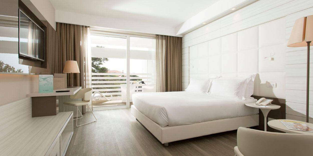 Hotel room with white bedding, wooden flooring, a neutral padded headboard and neutral lighting and cushions, with floor-to-ceiling windows overlooking a balcony