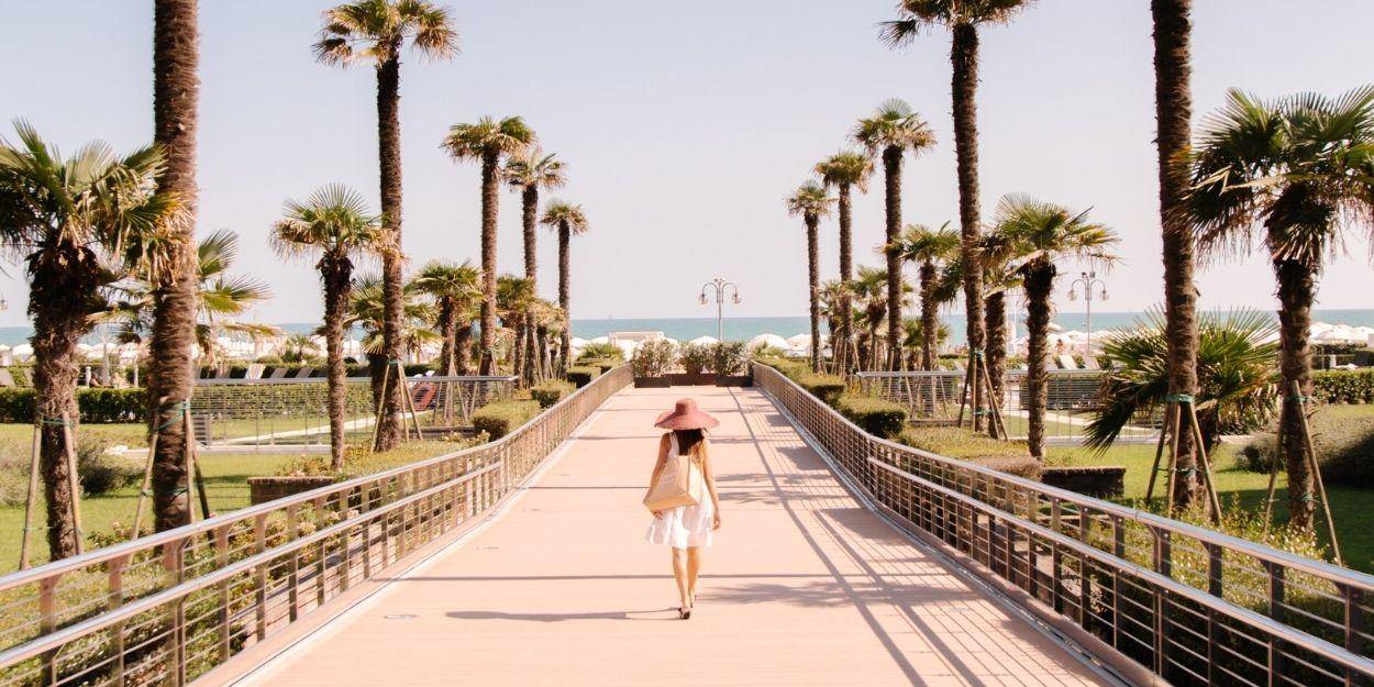 Woman in white dress and straw hat strolls across a paved bridge lined by palms towards the sea