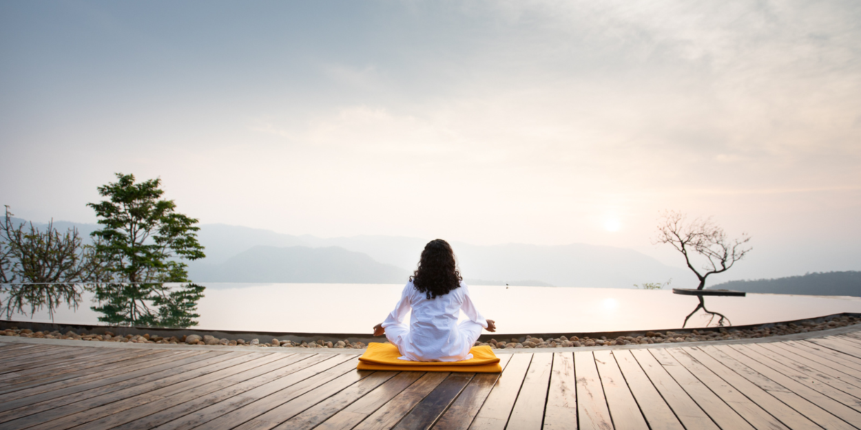 Woman sits on the edge of an infinity pool on a wooden deck meditating with misty hills in the distance