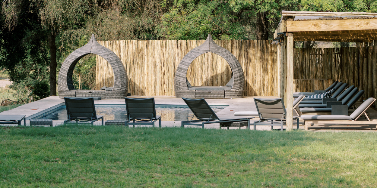 Rectangular swimming pool with striped cushion loungers and a wooden canopy, and rattan domed shelters next to a grassy lawn