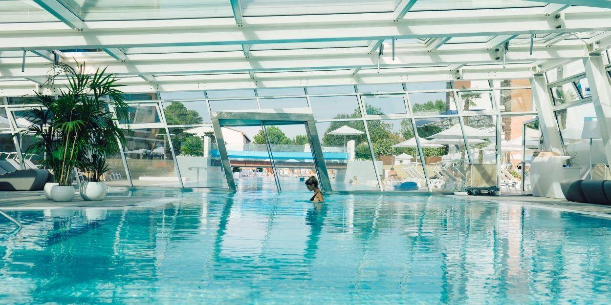Woman in a black swimsuit relax in an indoor pool that leads outdoors with wraparound glass windows and ceiling