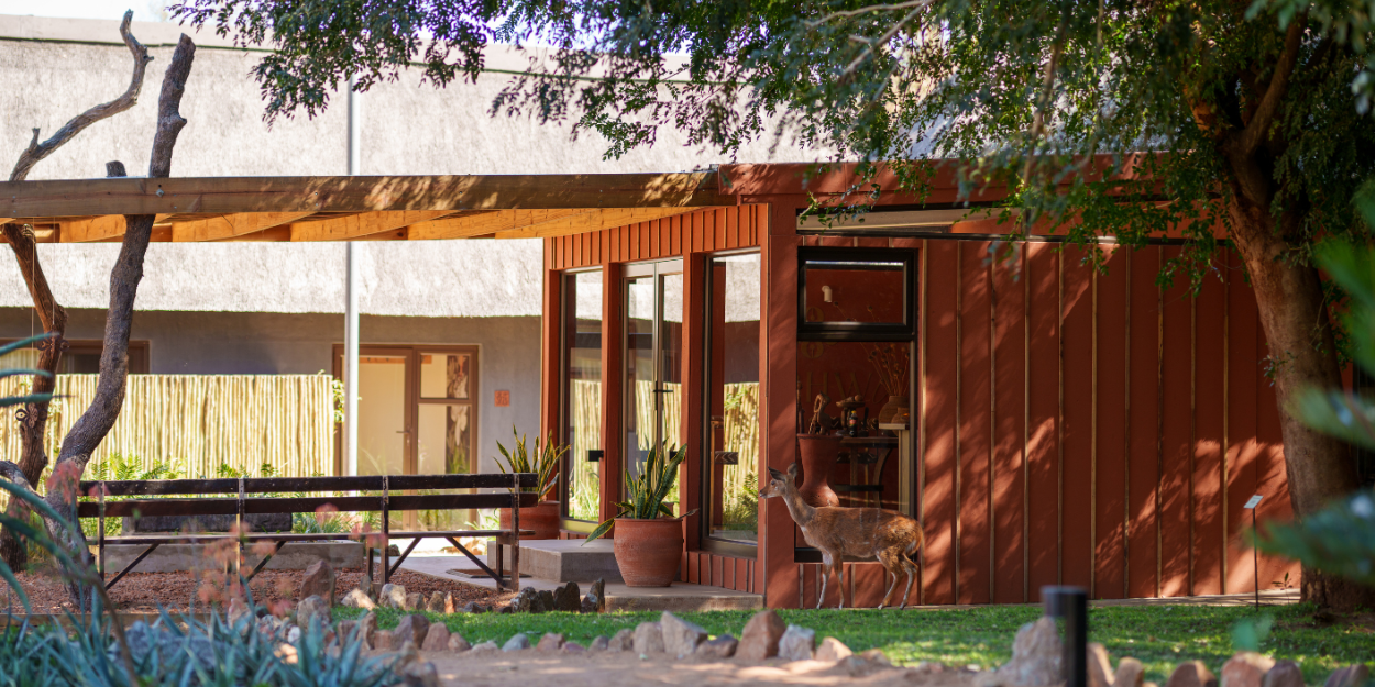 Low-rise terracotta building in a lawned area with trees, and a deer walking past