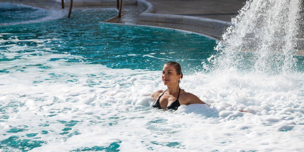 Woman in a dark swimsuit relaxes under a waterfall in a swimming pool