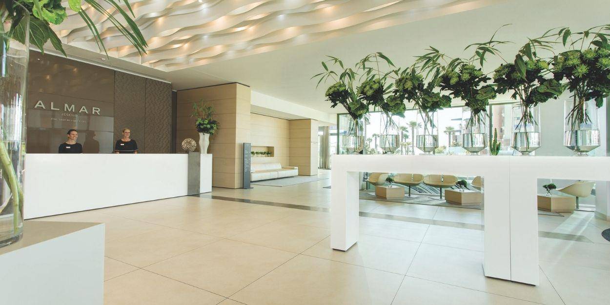 Smiling receptionists in a marble lobby with multiple vases of flowers, and a sign that reads 'Almar Jesolo Resort & Spa'