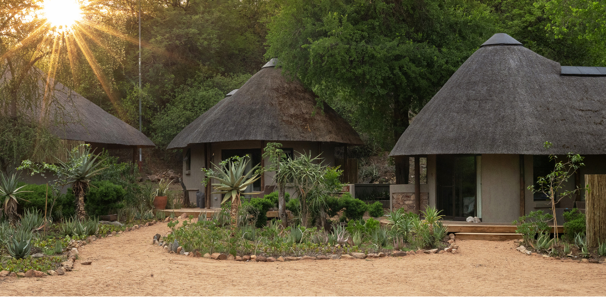 Triangular-roofed huts in a row surrounded by greenery as the sun dips behind the trees