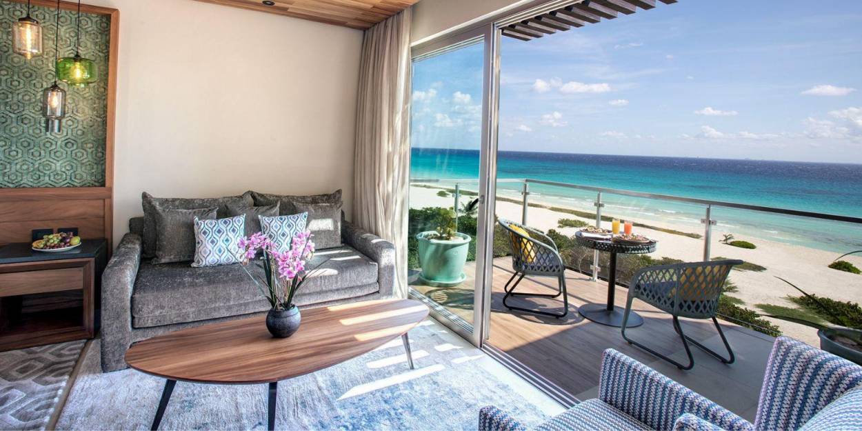 Hotel room living area at Palmaïa – The House of AïA Hotel room seating area with a grey sofa, blue rug and wooden coffee table, next to an open furnished balcony overlooking the beach