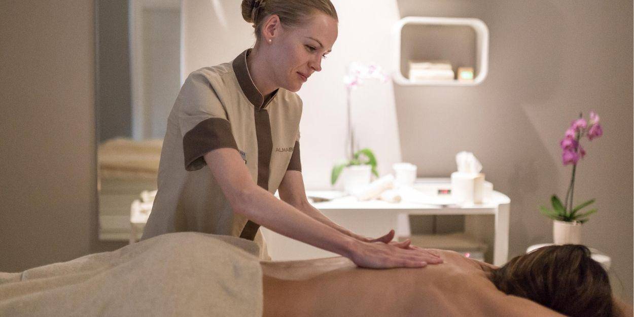 Smiling spa therapist massages a woman's back in a neutral treatment room with a pink potted flower and white vanity unit