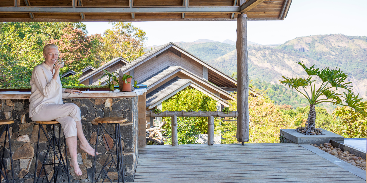 Smiling woman sips from a cup as she sits on an outdoor terrace overlooking green-cloaked mountains