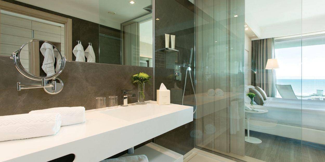 Sleek hotel bathroom with a marble wall, glass wall showing the bedroom, white long vanity unit and robes hanging up