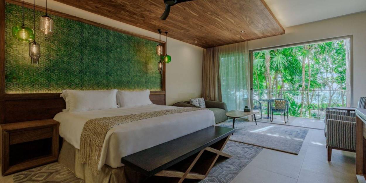 Accommodation at Palmaïa – The House of AïA Nature-inspired hotel room with neutral tones, an earth-green wall mural, a wooden ceiling and patio doors that open to a furnished balcony overlooking the mangroves