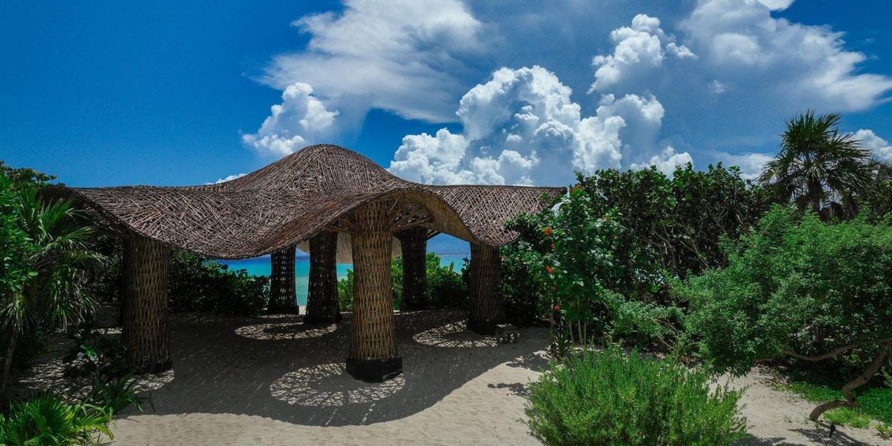 Outdoor space at Palmaïa – The House of AïA Pergola with wooden stilts and a curved thatched roof, surrounded by tropical greenery and overlooking the sea
