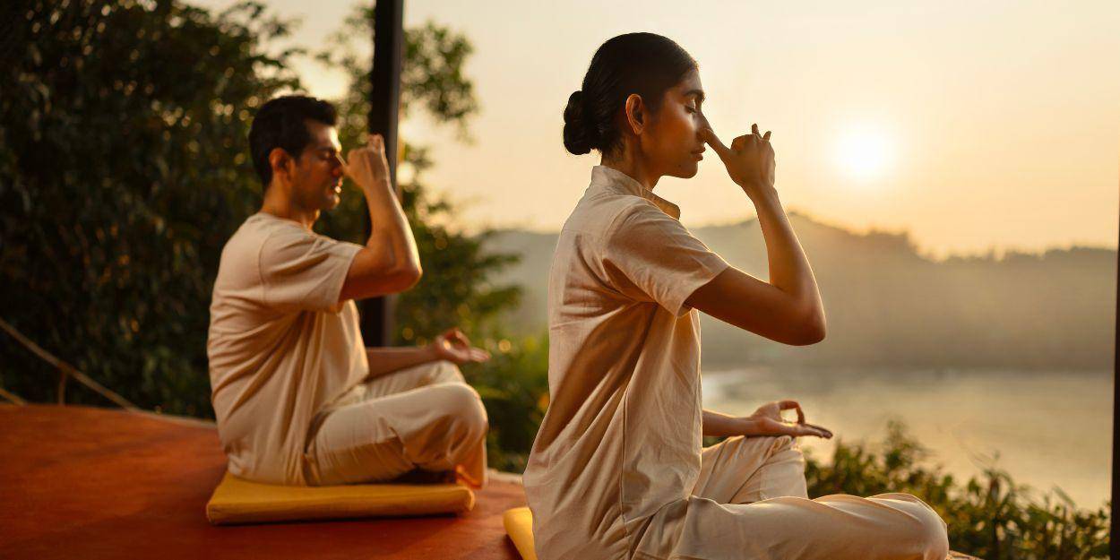 Two people in light, loose clothes sitting in yoga poses doing a breathwork on a wooden deck overlooking a curvy beach and the ocean