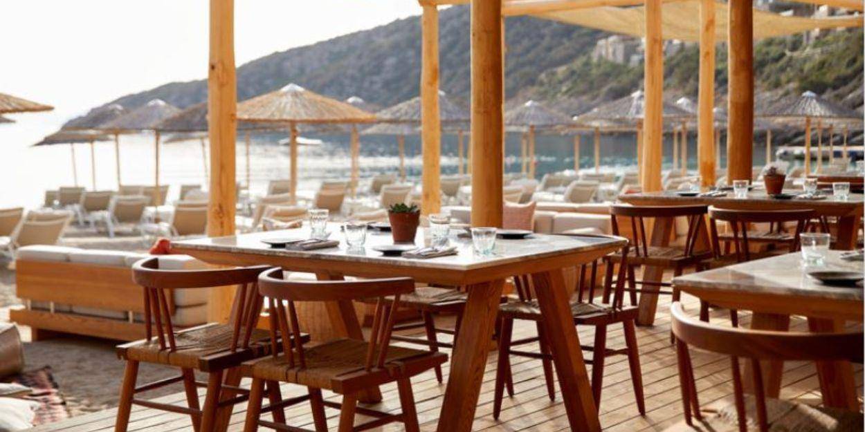 Beach club at Daios Cove Rustic-chic beach club with wooden flooring and stilts, a terrace with neutral sofas and thatched umbrellas in the background