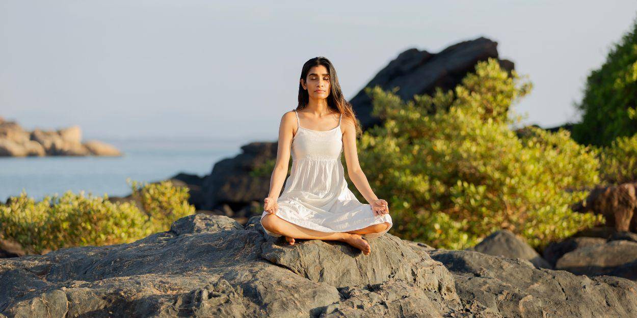 Woman sitting by the ocean practising meditation on rocks