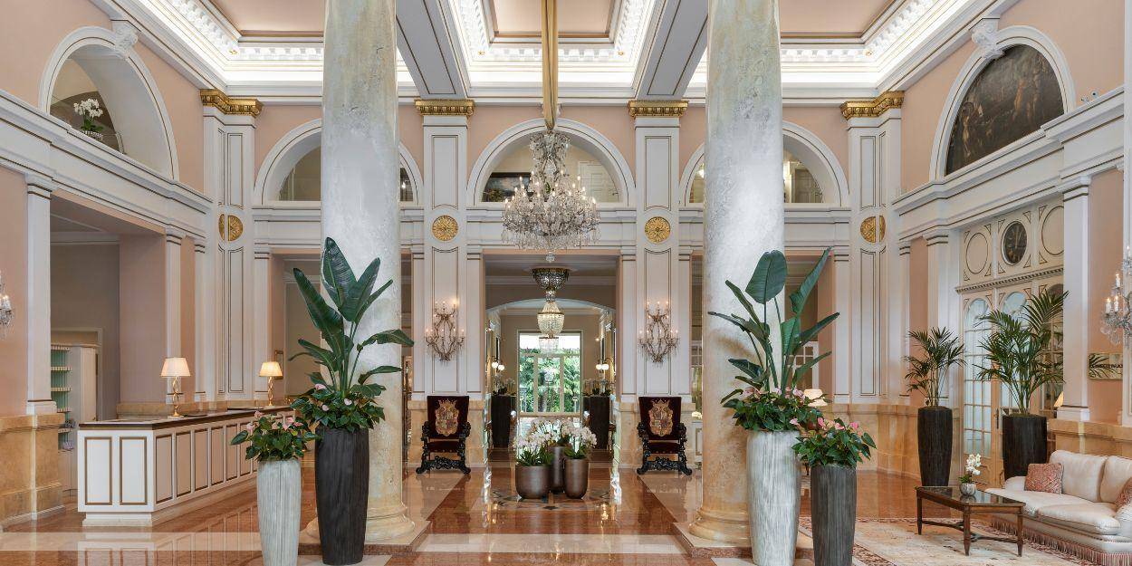 High ceilinged lobby area with ornate rugs and armchairs, gold accents, glittering chandelier-stye wall lights and grand white pillars
