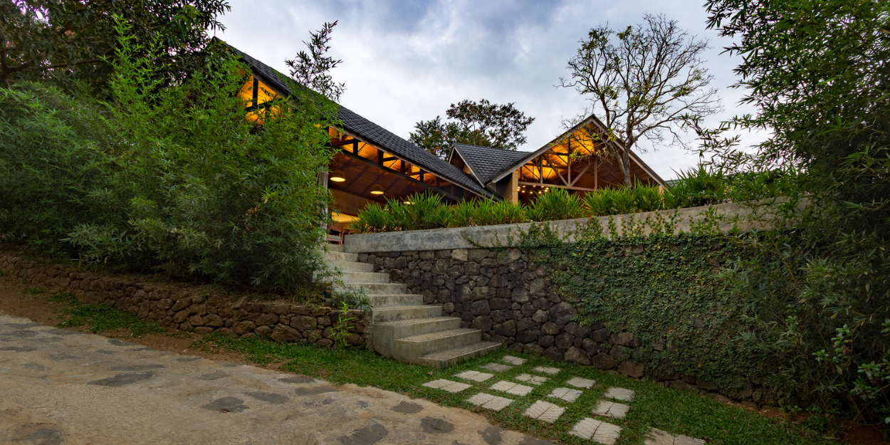 Stone pathways leading to a garden area and a building lit up at twilight, with stone and wood exteriors and a triangular roof
