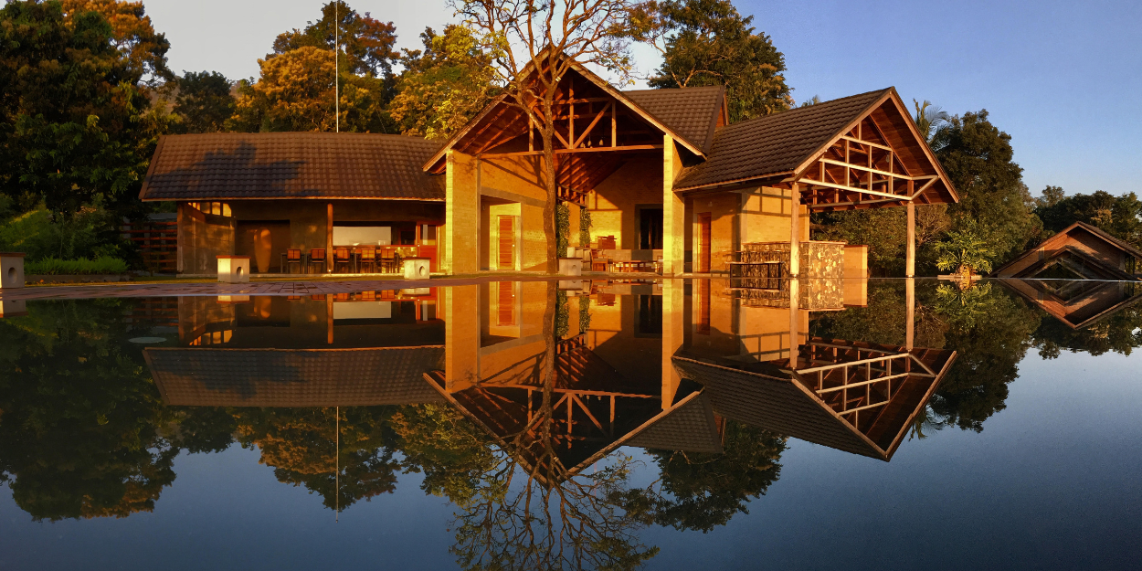 Lake-like swimming pool under a blue sky among tropical greenery, overlooked by a wooden building with triangular rooftops and stilts