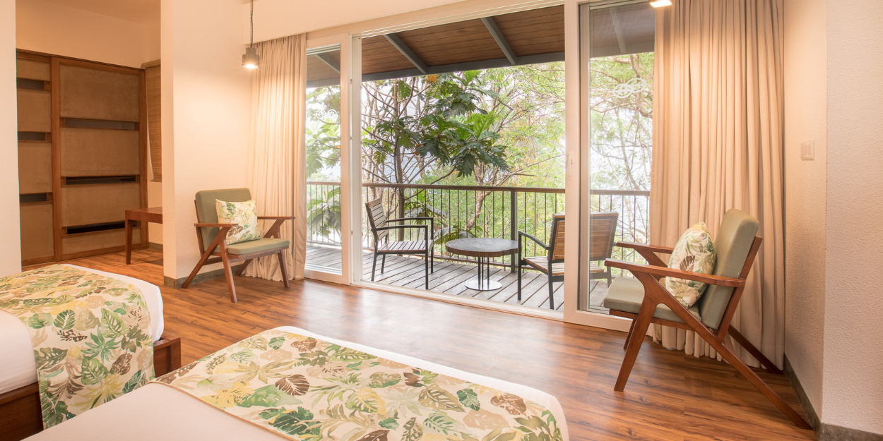 Hotel room with wooden floors and wardrobes, white walls and bedding, green soft furnishings and patio doors open onto a furnished balcony