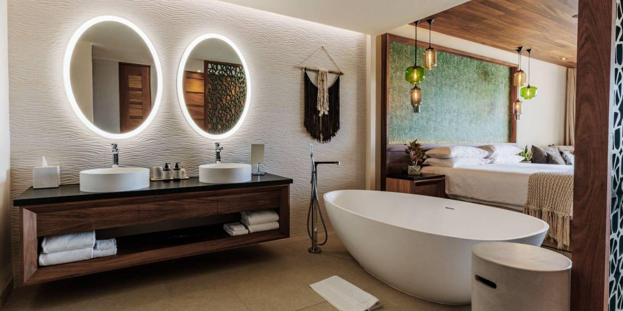Accommodation at Palmaïa – The House of AïA Bathroom with a large window overlooking the bedroom, with twin sinks, mirrors, a standalone bathtub, and dark-wood furnishings with a macrame design on the wall