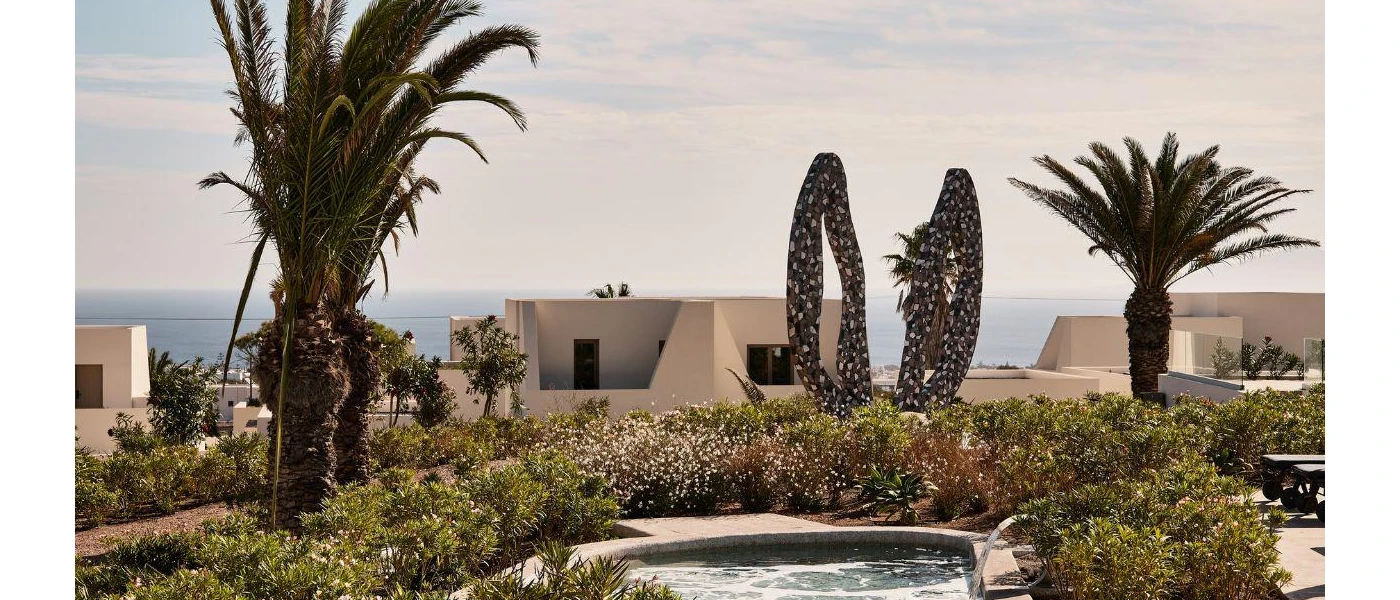 Pond with water jets surrounded by bushes, palm trees and a donkey ears sculpture, with low-rise cube-like accommodation blocks and the sea in the background