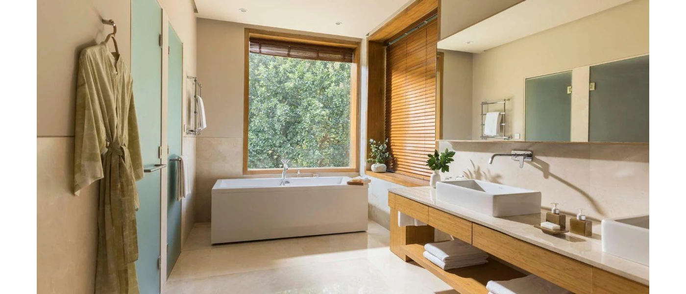 Neutral bathroom with wooden furniture and wall panels, a white standalone bathtub next to a large window, his and hers white sinks and a beige robe hanging on a hook, 