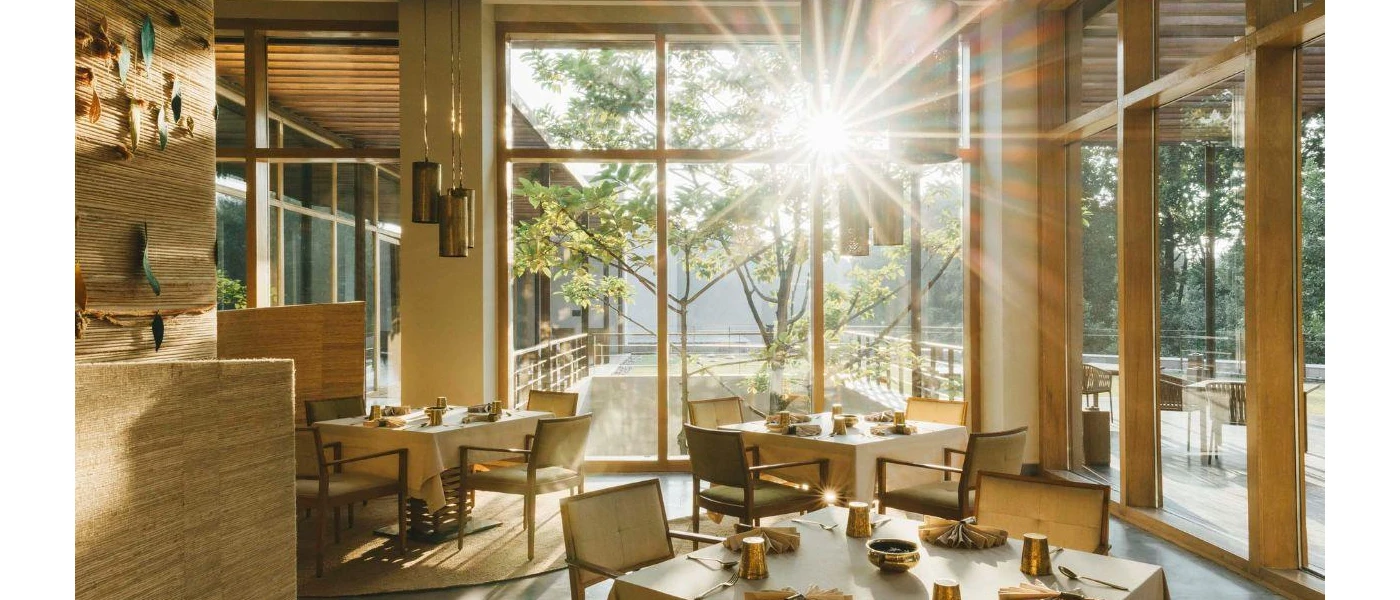 Airy restaurant with sun rays beaming through floor-to-ceiling windows, textured rattan-style walls and neutral table set-ups with gold cups