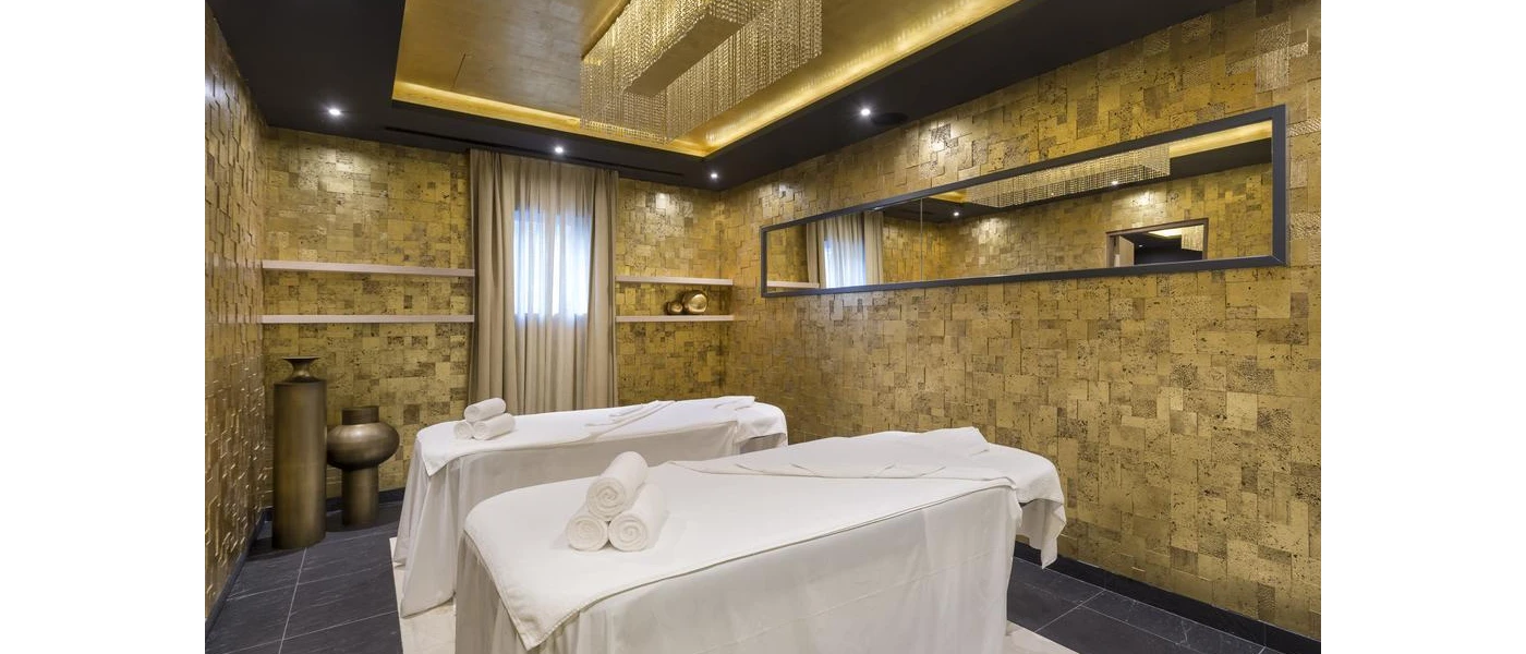 Treatment room with a gold chandelier and decorative pots, tiled walls and two massage beds covered in white towels