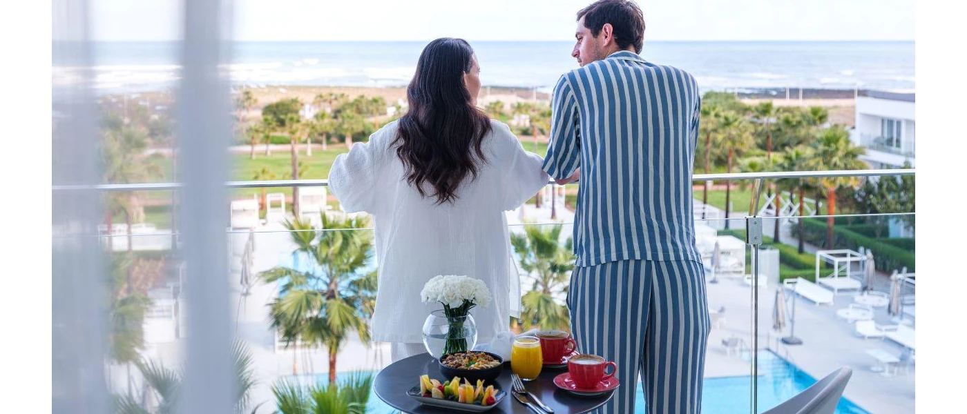 Dark-haired couple in pyjamas standing on a glass balcony looking down on grounds covered in palm trees and a central swimming pool