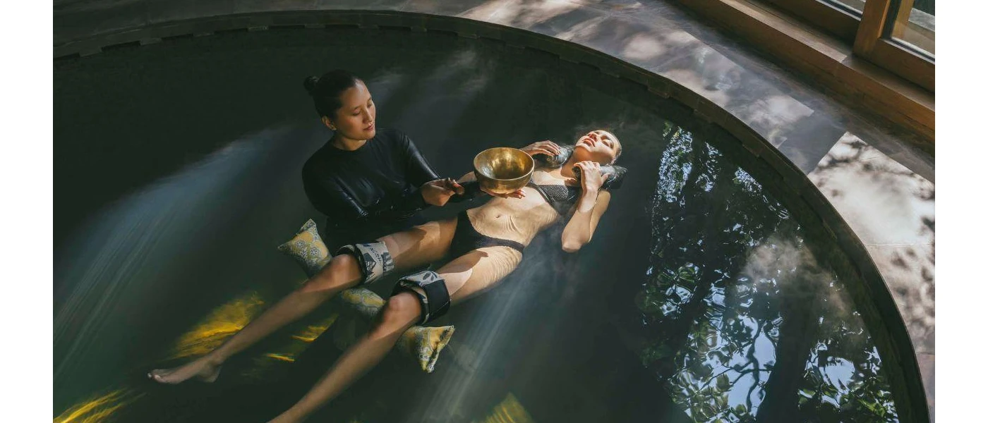 Woman in a black bikini lies with weights in a floatation pool, while a therapist supports her body and and plays a sound bowl