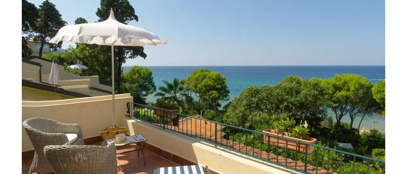 Sunny terrace with a white parasol and grey wicker chairs overlooking trees and the sea