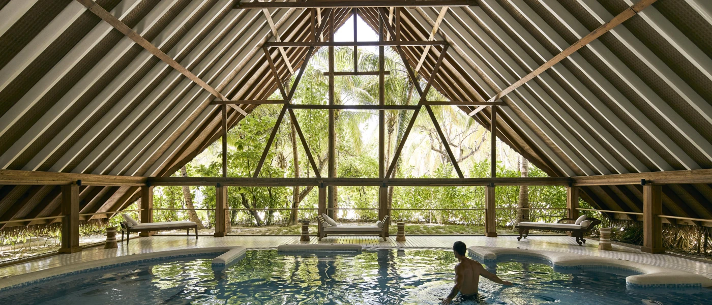 Indoor pool with jets in a room with a triangular wood-beamed roof and floor-to-ceiling windows with a view of tropical gardens