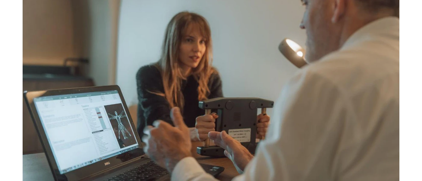 Staff member in a white coat explains findings on a screen to a woman holding a digital device