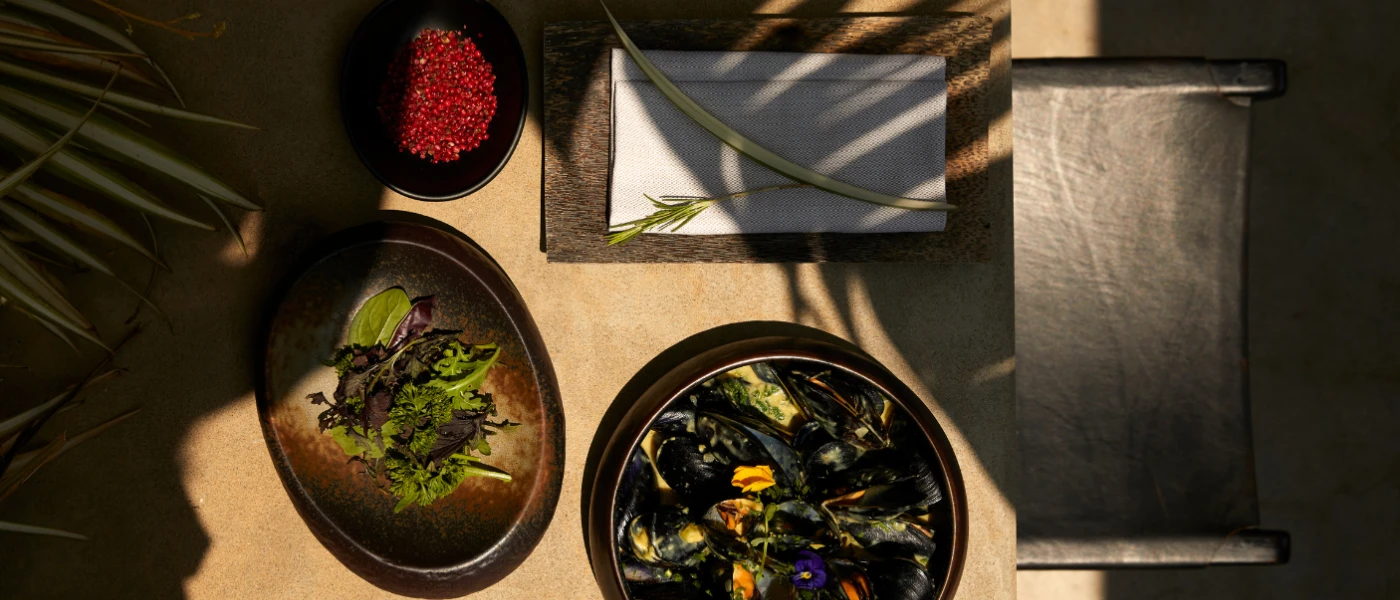Table laid up with a bowl of steamed mussels, a plate of pomegranates and a plate of salad under the shade of palm trees
