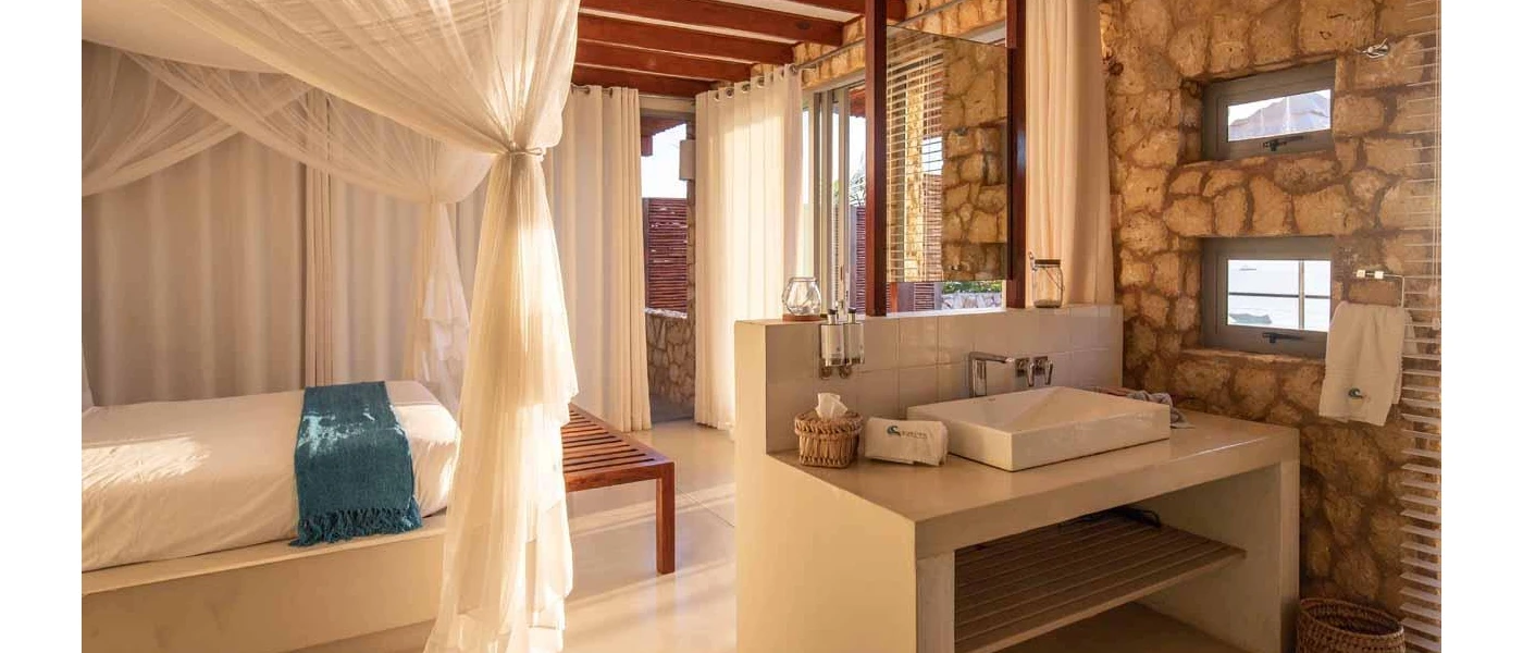 Hotel room with stone walls, a beige vanity unit with white sink and four-poster bed with white drapes
