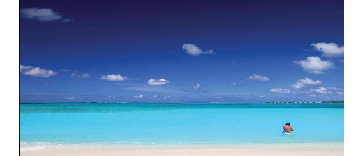 Man stands in bright turquoise shallows with a gleaming white beach behind, under a blue sky