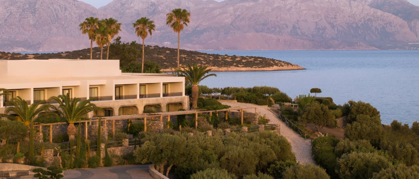 White two-storey building with cobbled stone walls, surrounded by stone pathways, greenery and hills and the sea