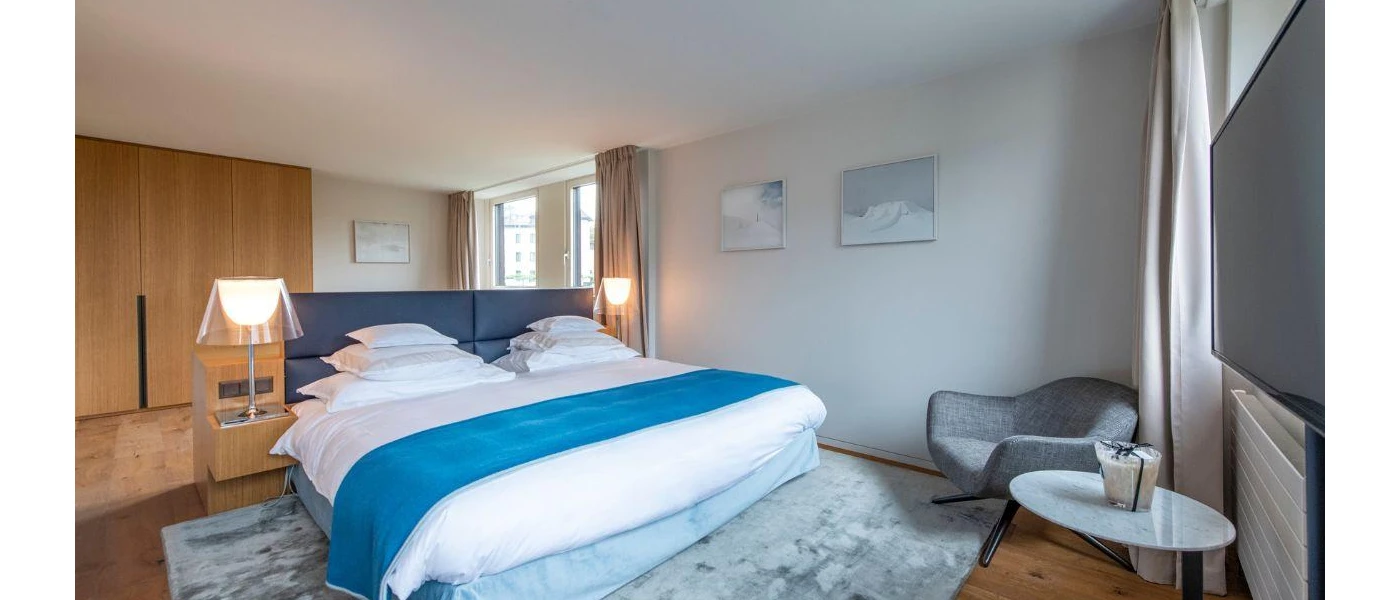 Modern hotel bedroom with crisp white bedding with a blue headboard and throw, a grey patterned rug underneath, light-wood wardrobe, a grey corner chair and table and a wall-hung TV