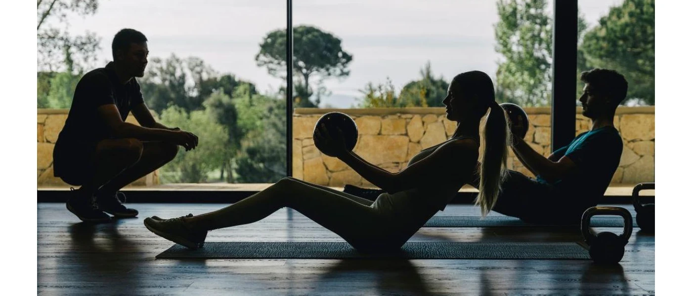 Male and female work out with medicine balls on mats in a studio with floor-to-ceiling windows, as a trainer watches from the front