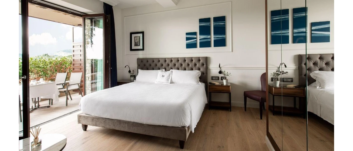 Modern hotel room with glass-door wardrobes, white bedding, a grey padded headboard and patio doors open to a furnished terrace